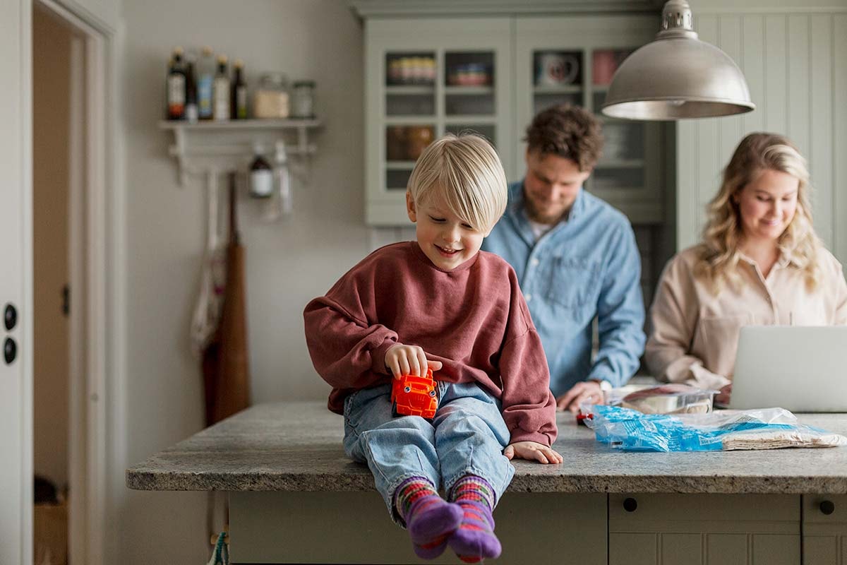 Gutt sitter på kjøkkenbenken og leker med bil med to voksne bak seg