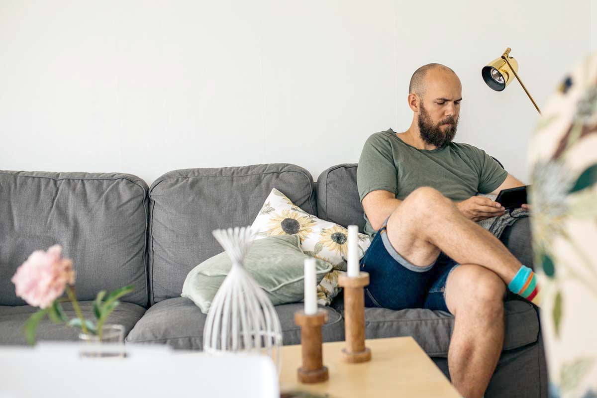 Mann sitter i sofa i eget hjem og leser på nettbrett, til illustrasjon for gjeldsregisteret
