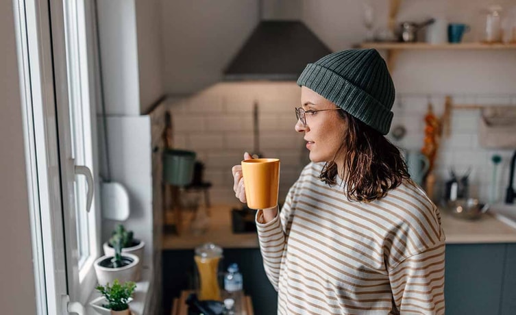 Dame med lue, briller og stripete genser står på kjøkkenet og drikker kaffe