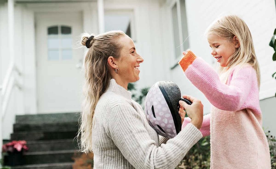 Kvinne står sammen med jente utenfor hjemmet og kler på jenta en hjelm, til illustrasjon for rammelån