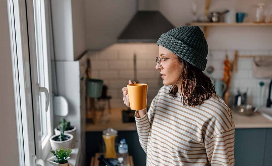 Dame med lue, briller og stripete genser står på kjøkkenet og drikker kaffe