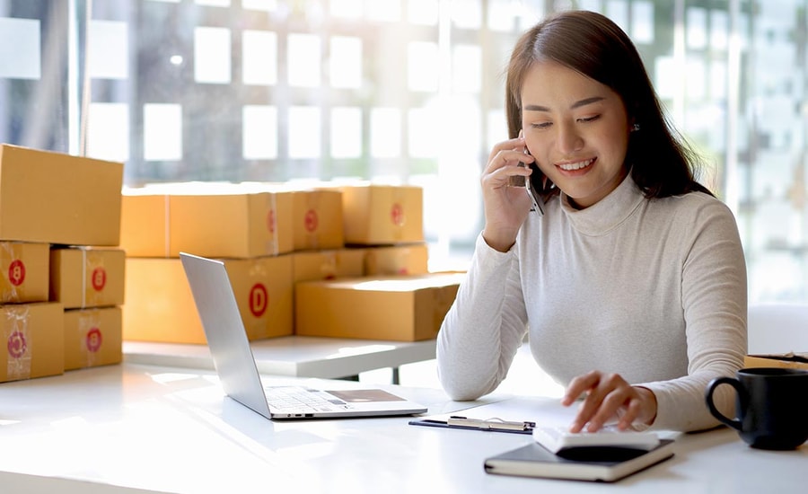 Dame sitter og snakker i telefon med PC ved siden av og mange bokser bak henne, til illustrasjon for avtalegiro