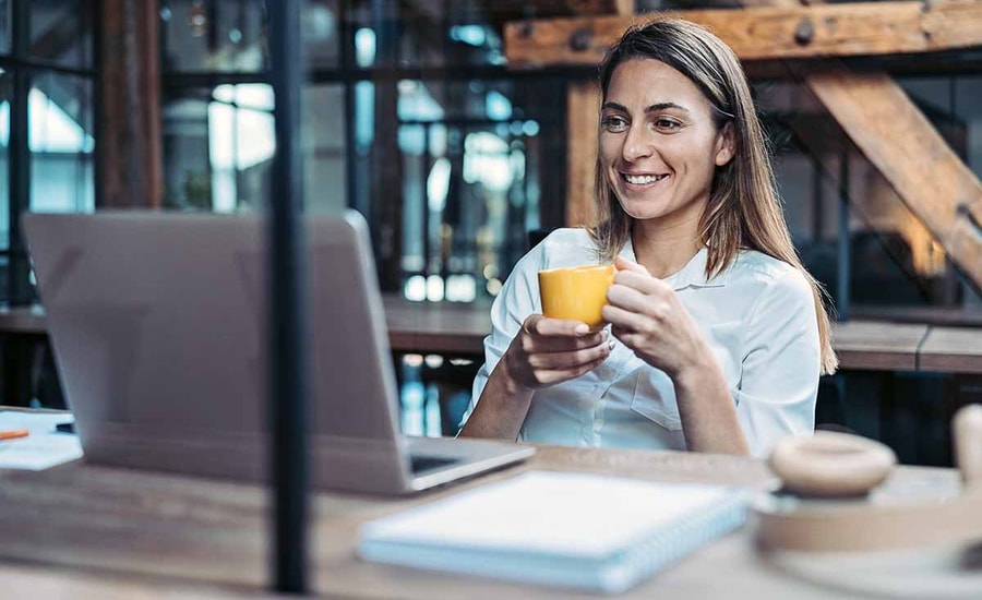 Dame sitter ved pult og ser på mac mens hun holder en kopp, til illustrasjon for faktura med kid