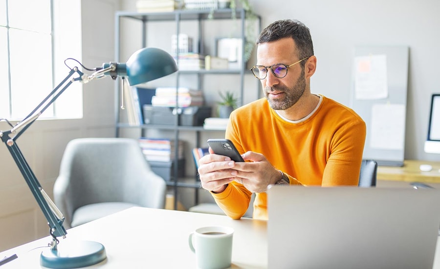 Mann i gul genser sitter ved et skrivebord med en kopp kaffe og bruker mobilen og PC, til illustrasjon for identitetstyveri