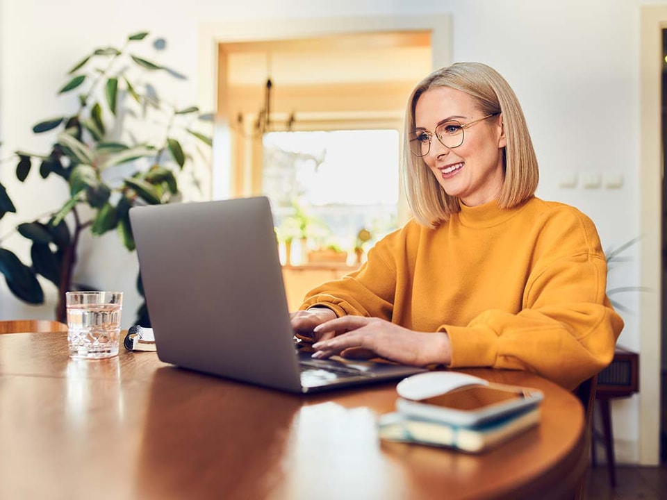 Dame i oransje grenser sitter ved spisebord og ser på datamaskin, til illustrasjon for betale til utlandet