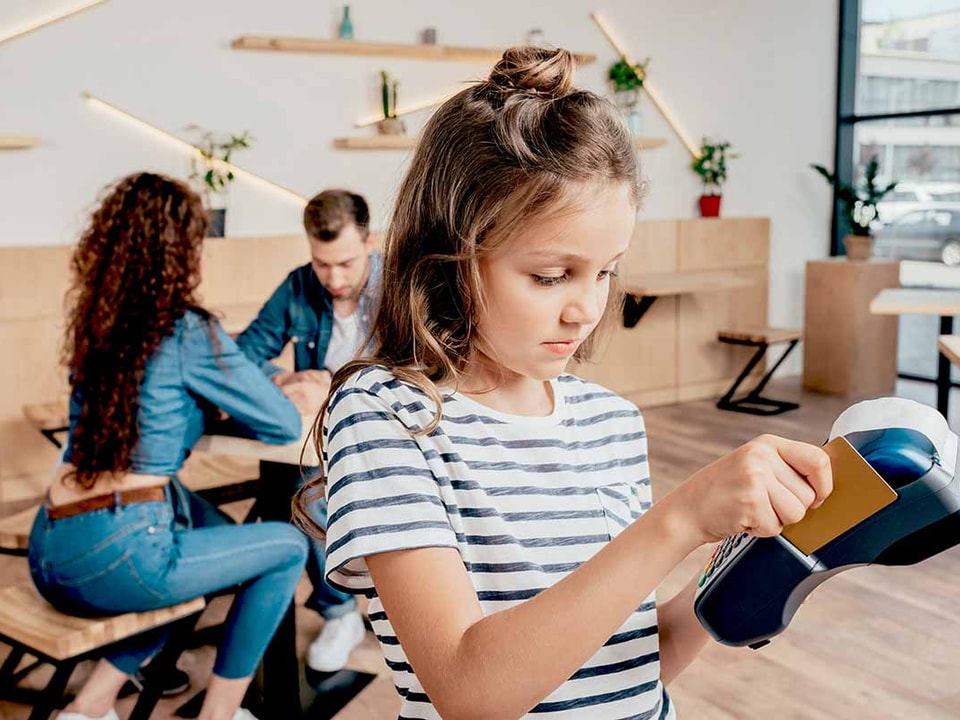 Ung jente betaler med kort på betalingsterminal med to voksne i bakgrunnen, til illustrasjon for barn, penger og økonomi