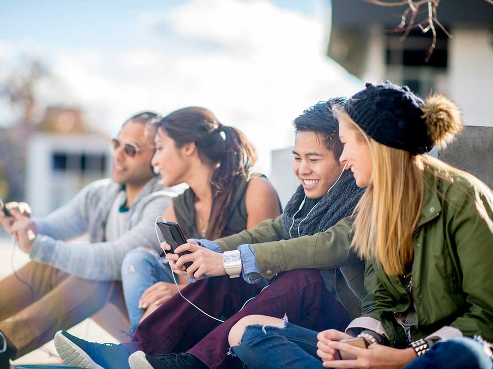 Ungdommer sitter på bakken og ser på mobilene sammen, til illustrasjon for bank for barn og ungdom