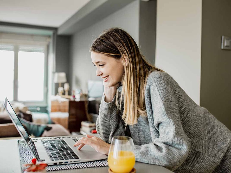 Dame jobber på PC over kjøkkenbenken med et glass juice, til illustrasjon for dagligbank
