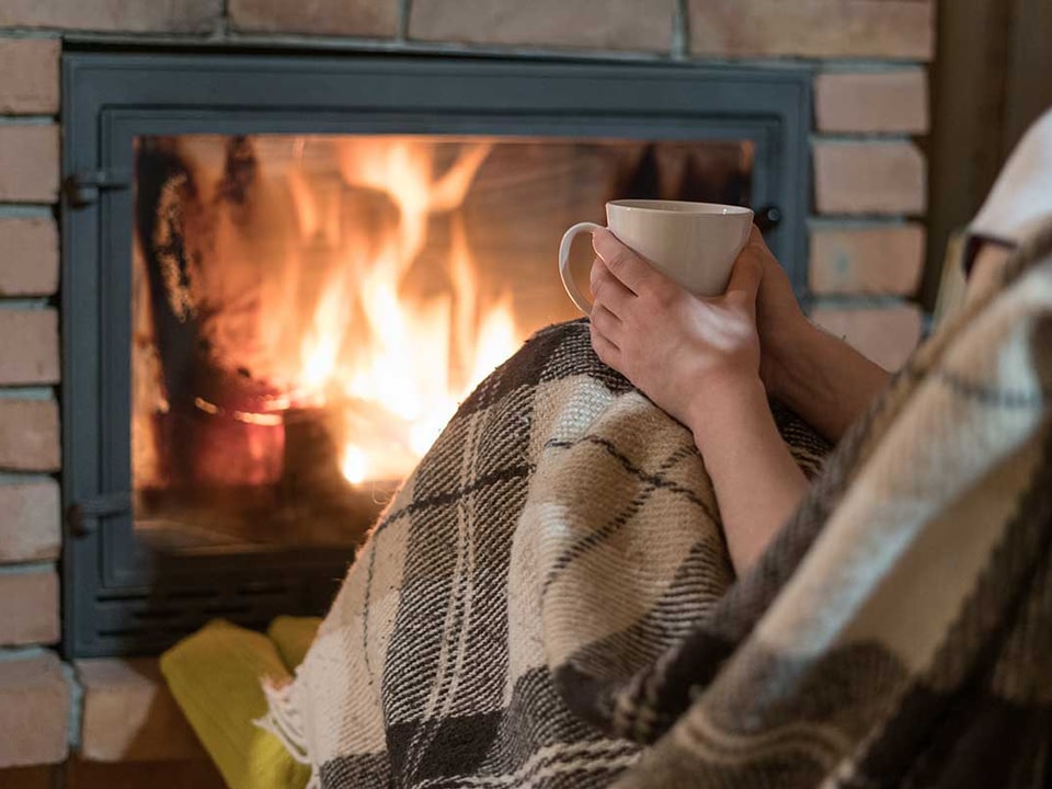 Dame sitter foran peis med en kopp i handa, til illustrasjon for energieffektivisere bolig