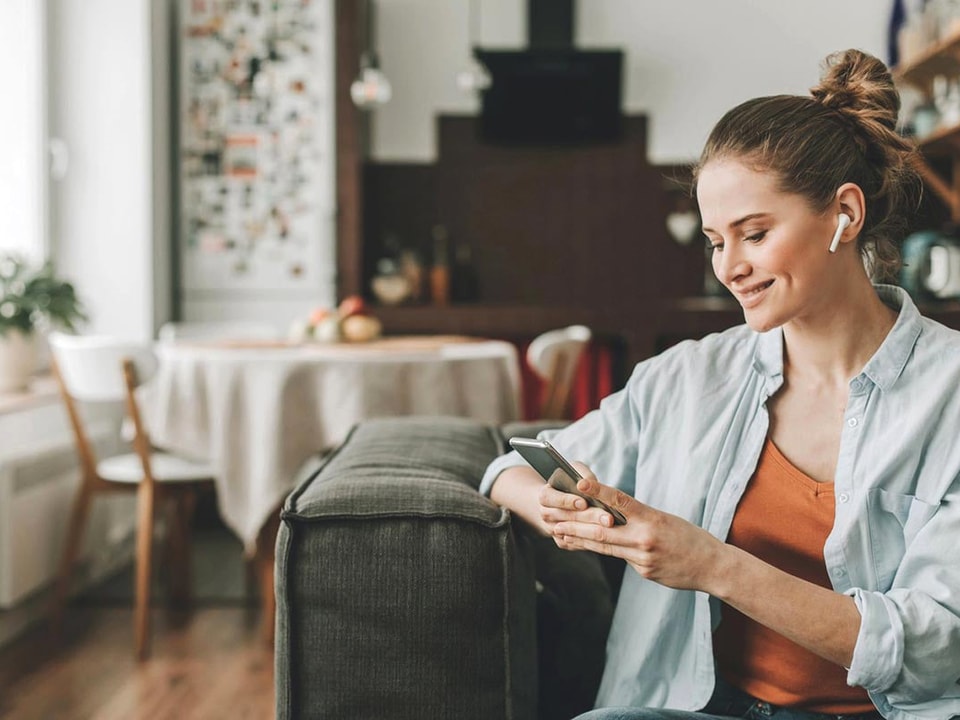 Dame sitter i en sofa med airpods i ørene og skriver på mobiltelefon, til illustrasjon for eiendomsmegler