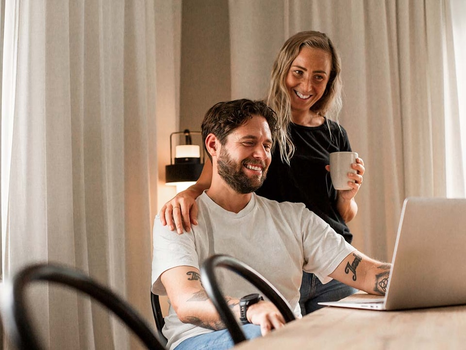 Kvinne holder armen sin rundt en mann som sitter og jobber på PC, til illustrasjon for høyrentekonto