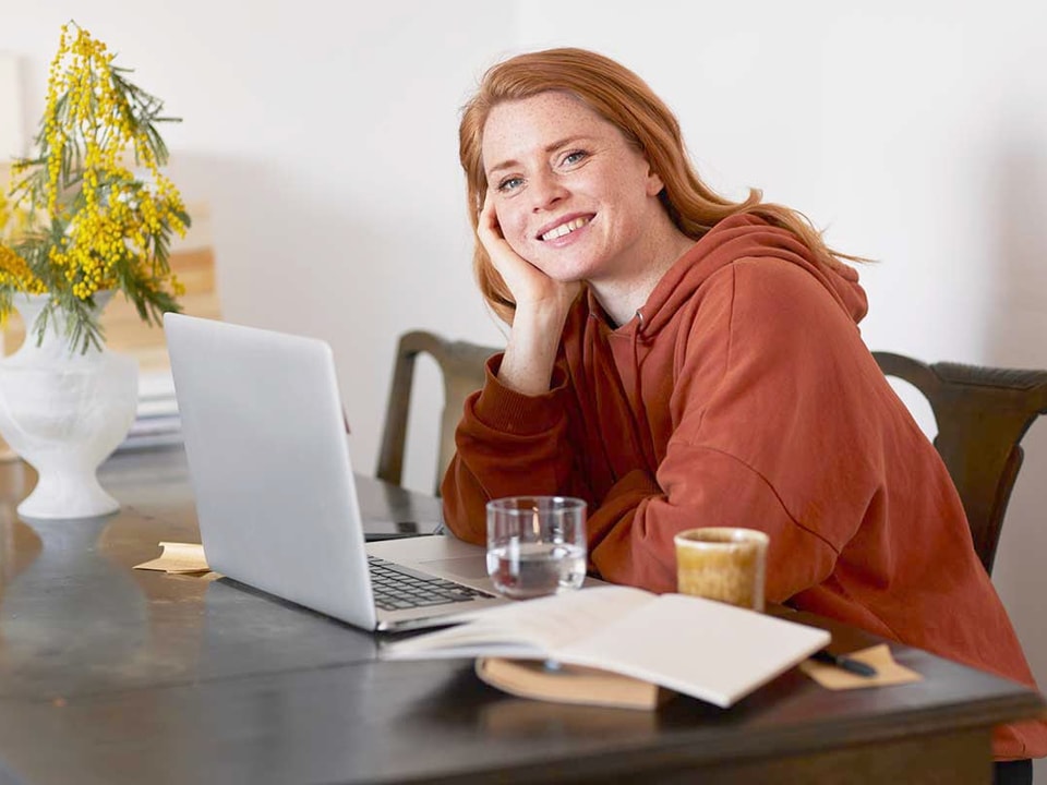 Ung jente sitter på kjøkkenbord med datamaskin fremfor seg og smiler, til illustrasjon for sparekalkulator