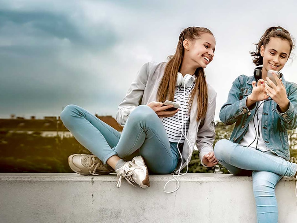 To tenåringstjenster med headset rundt halsen og mobilen i handa, til illustrasjon for sparekonto barn og ungdom