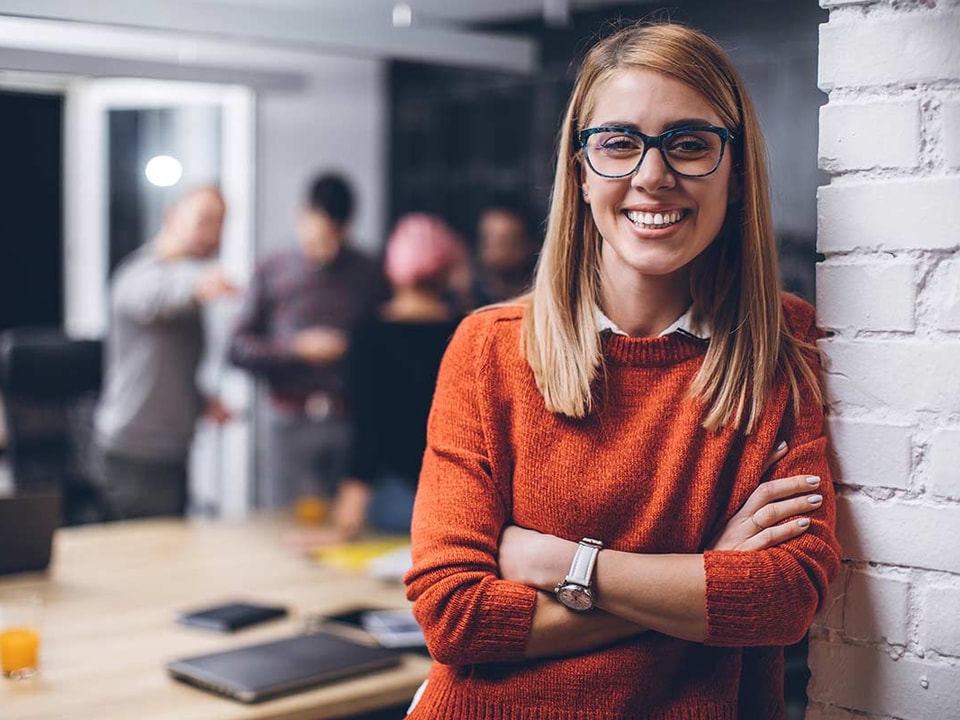 Kvinne med orange genser og briller smiler i kontorlokale, til illustrasjon for bli bedriftskunde