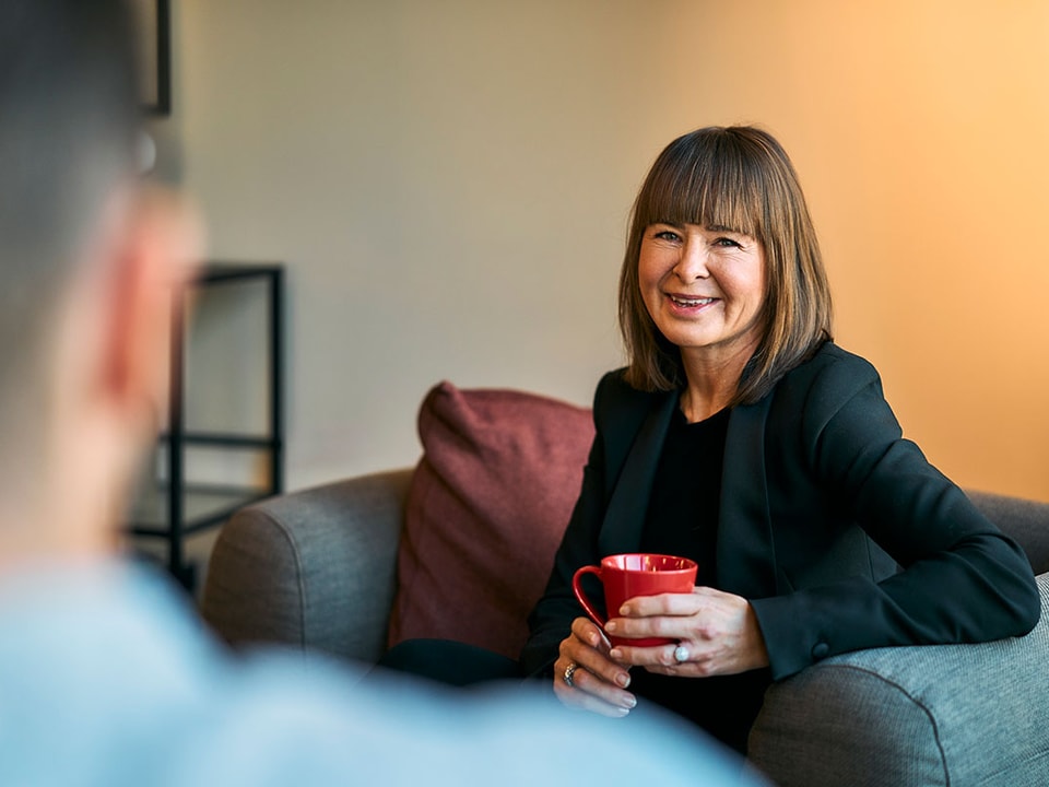Kvinne sitter i en grå stol og holder en rød kopp mens hun smiler, til illustrasjon for BN Bank