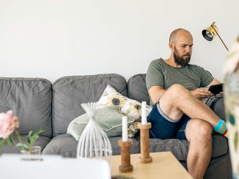 Mann sitter i sofa i eget hjem og leser på nettbrett, til illustrasjon for gjeldsregisteret