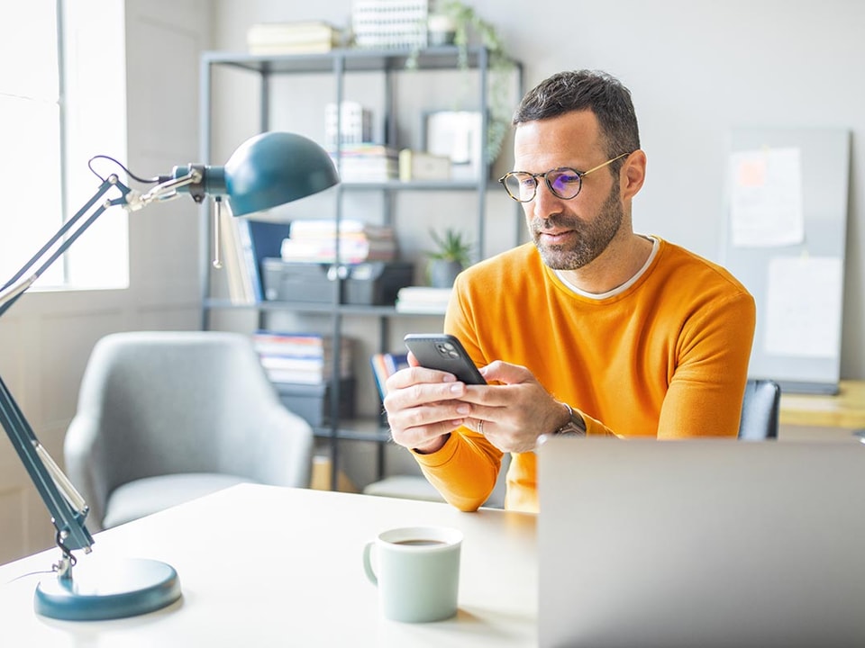 Mann i gul genser sitter ved et skrivebord med en kopp kaffe og bruker mobilen og PC, til illustrasjon for identitetstyveri
