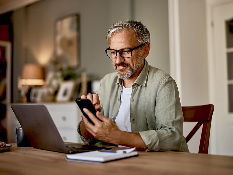 Mann med pc og som ser på mobilen, illustrasjon til nye sikkerhetskrav ved netthandel
