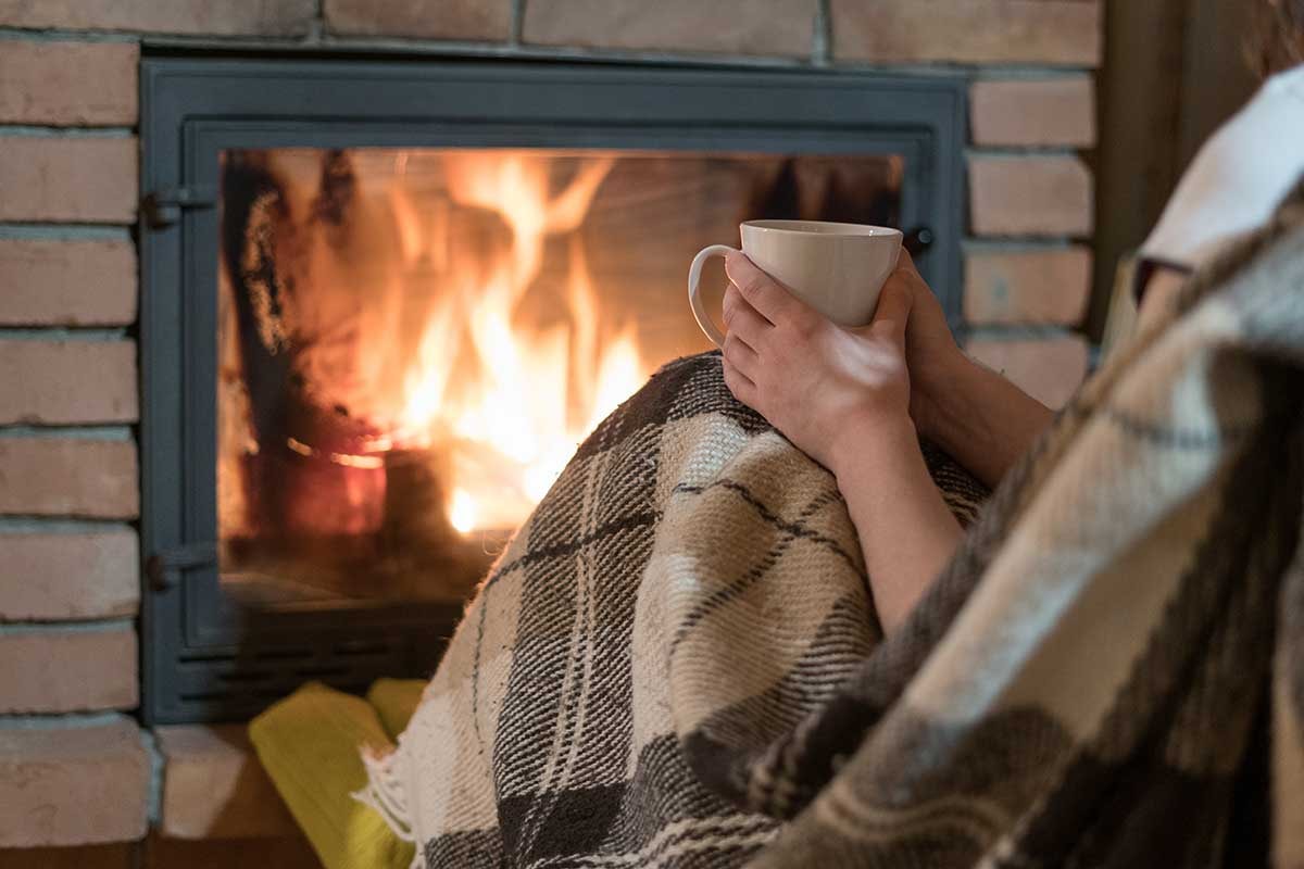 Dame sitter foran peis med en kopp i handa, til illustrasjon for energieffektivisere bolig