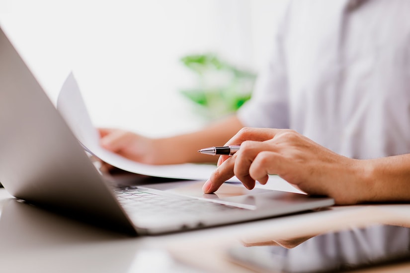 Person sitter med laptop og holder dokumenter og penn, til illustrasjon for avtalegiro