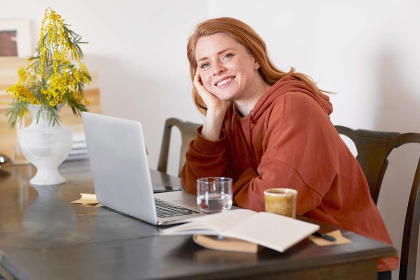 Ung jente sitter på kjøkkenbord med datamaskin fremfor seg og smiler, til illustrasjon for sparekalkulator