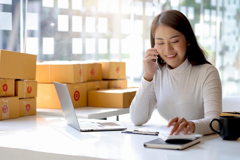Dame sitter og snakker i telefon med PC ved siden av og mange bokser bak henne, til illustrasjon for avtalegiro