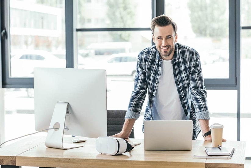Mann står ve pulten med mac og laptop, til illustrasjon for kapitalkonto