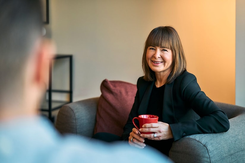 Kvinne sitter i en grå stol og holder en rød kopp mens hun smiler, til illustrasjon for BN Bank