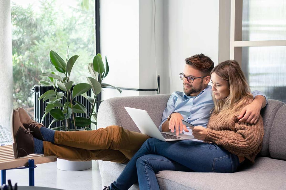 Par sitter i sofaen og ser på laptop, til illustrasjon for omkostninger ved boligkjøp