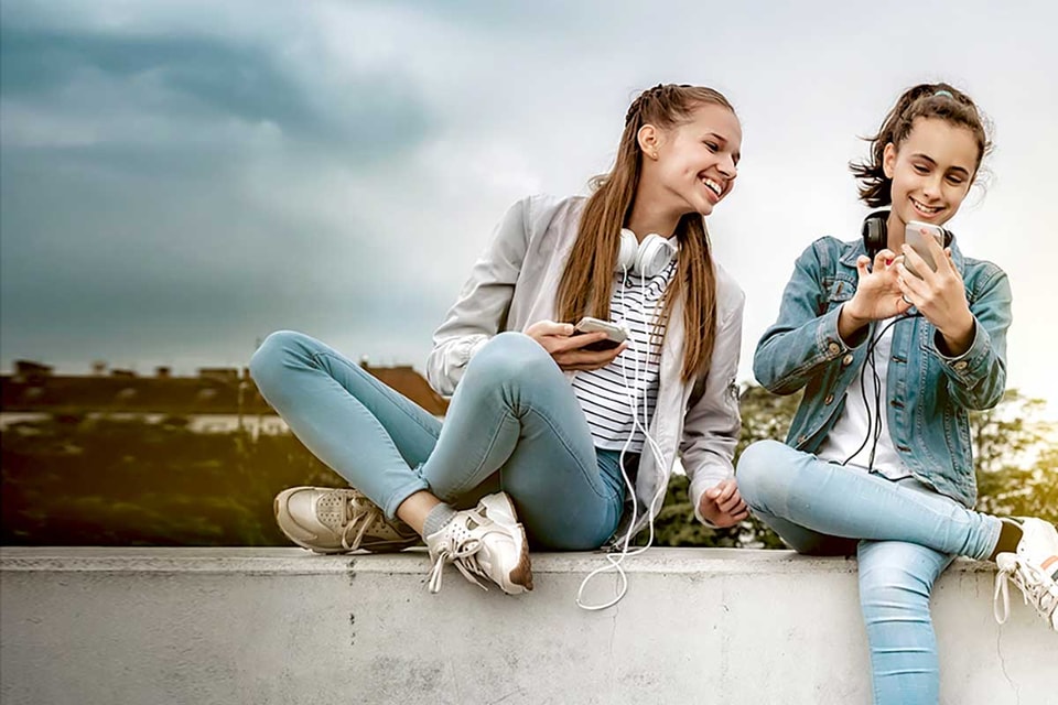 To tenåringstjenster med headset rundt halsen og mobilen i handa, til illustrasjon for sparekonto barn og ungdom