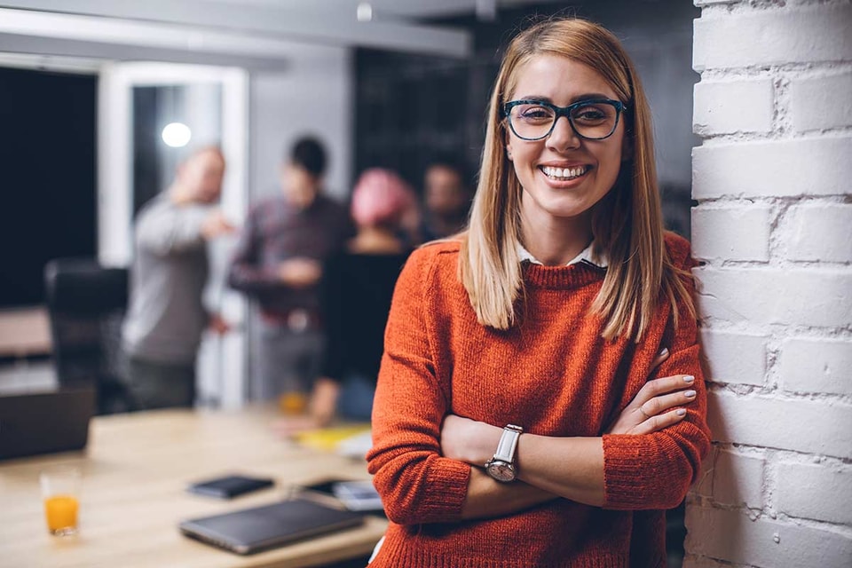 Kvinne med orange genser og briller smiler i kontorlokale, til illustrasjon for bli bedriftskunde