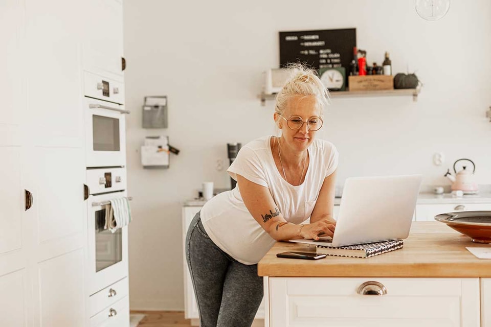 Dame med laptop står ved kjøkkenbenken, illustrasjon til veileder om hvordan legge til disponent på konto