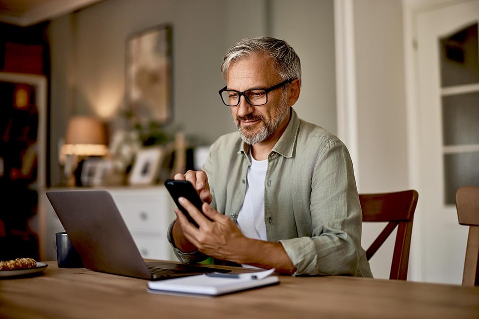 Mann med pc og som ser på mobilen, illustrasjon til nye sikkerhetskrav ved netthandel