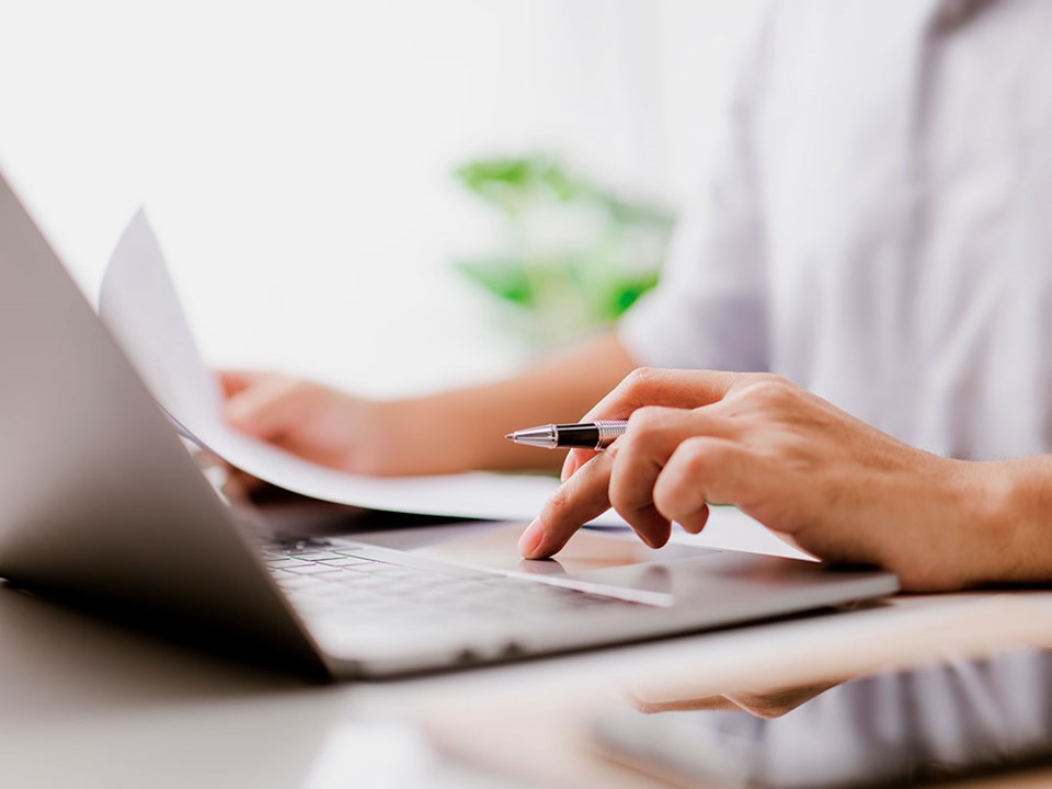 Person sitter med laptop og holder dokumenter og penn, til illustrasjon for avtalegiro