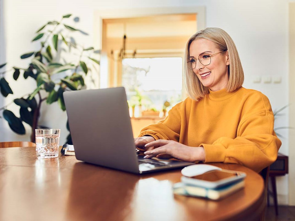 Dame i oransje grenser sitter ved spisebord og ser på datamaskin, til illustrasjon for betale til utlandet