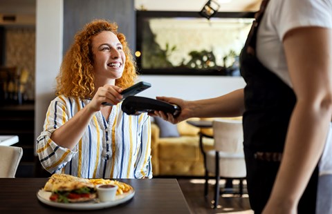 Dame betaler med mobiltelefonen på restaurant og smiler, til illustrasjon for Samsung Pay