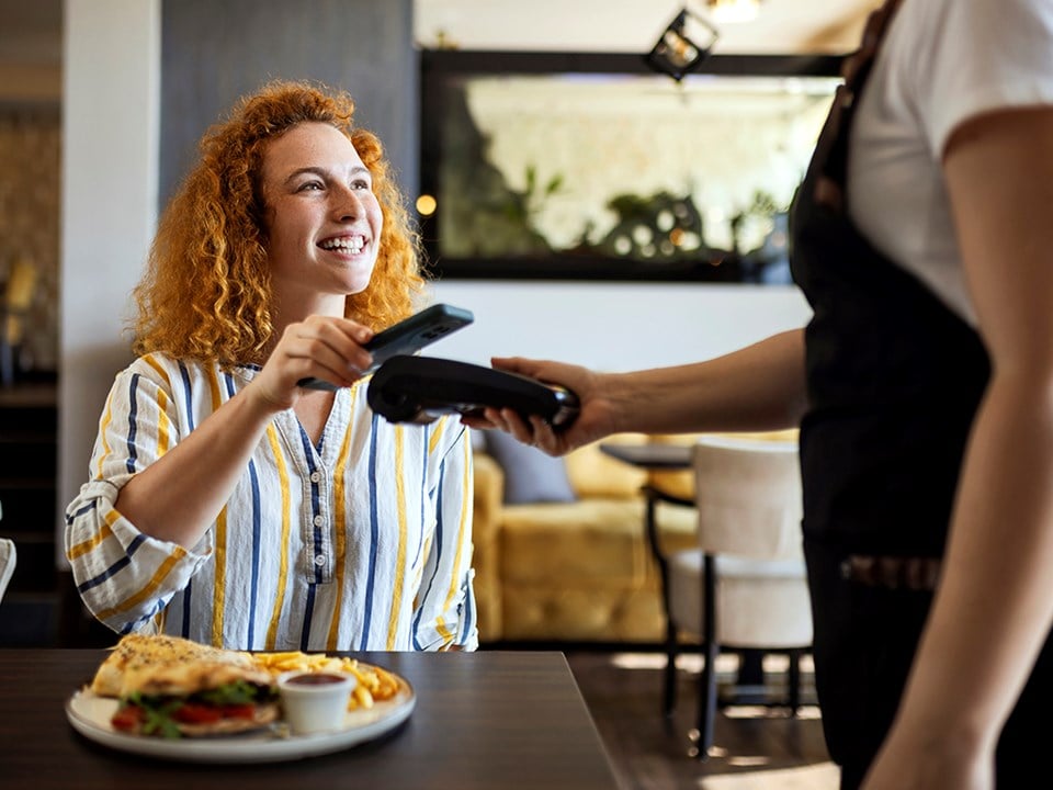 Dame betaler med mobiltelefonen på restaurant og smiler, til illustrasjon for Samsung Pay