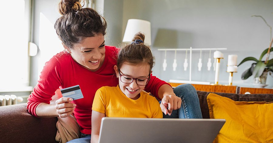 Mor og barn sitter på sofa og ser på datamaskin med bankkort i handa, til illustrasjon og barn og penger
