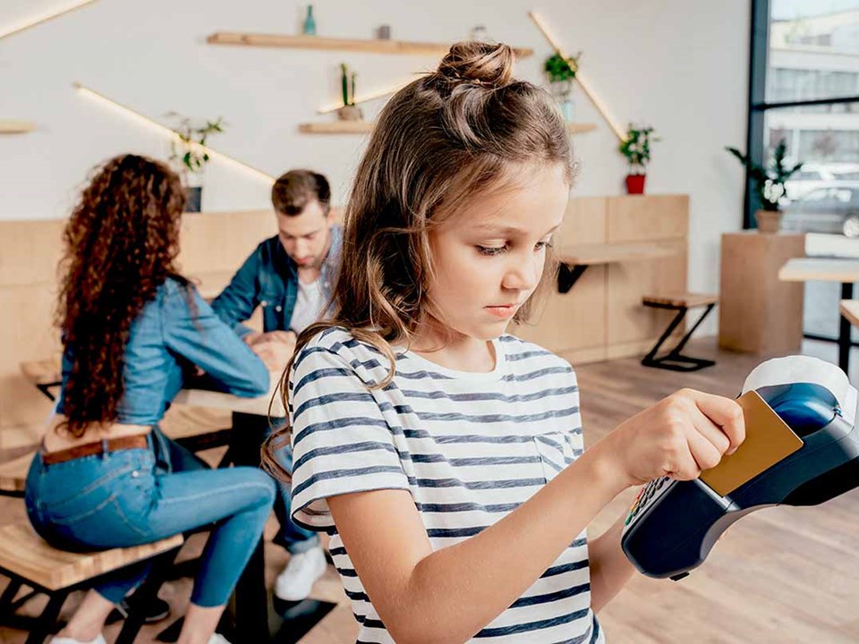 Ung jente betaler med kort på betalingsterminal med to voksne i bakgrunnen, til illustrasjon for barn, penger og økonomi