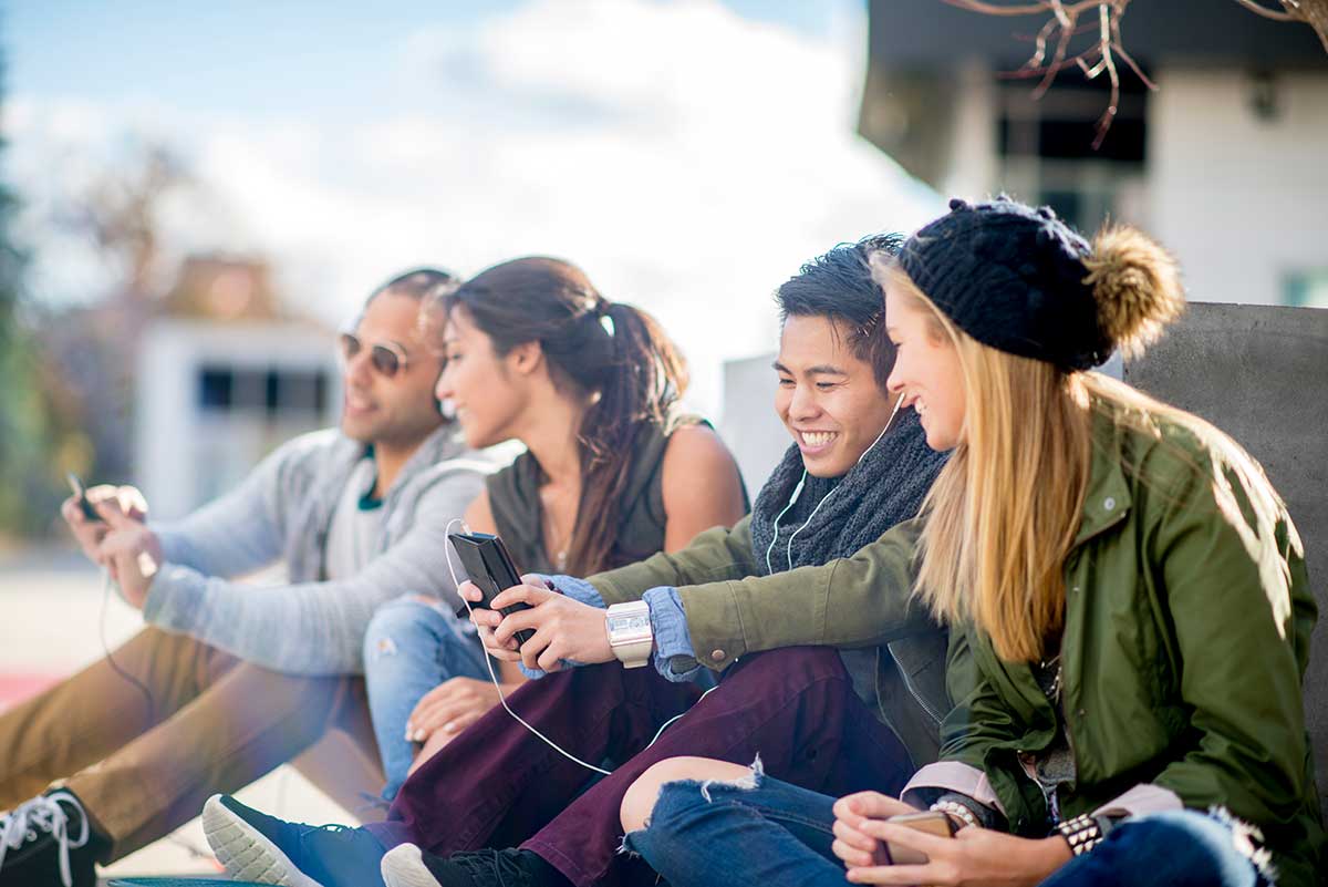Ungdommer sitter på bakken og ser på mobilene sammen, til illustrasjon for bank for barn og ungdom