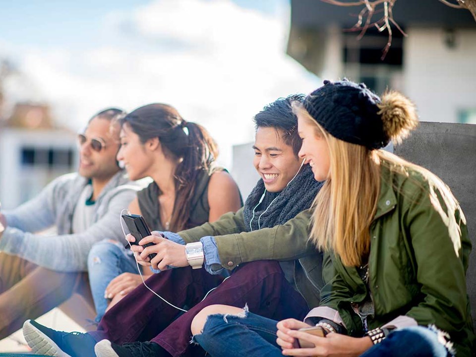 Ungdommer sitter på bakken og ser på mobilene sammen, til illustrasjon for bank for barn og ungdom