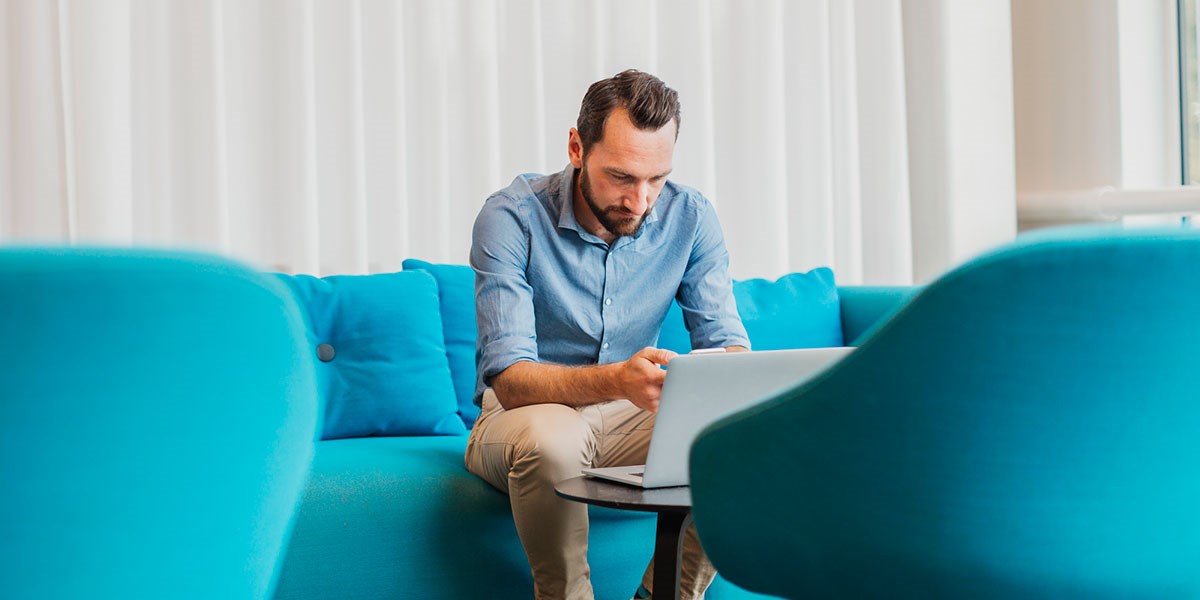 Mann sitter i blå sofa og jobber på PC, til illustrasjon for trygg kortbruk