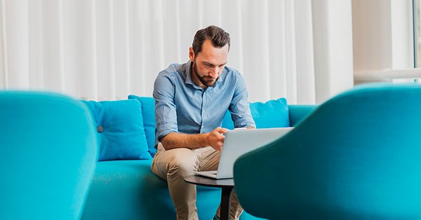 Mann sitter i blå sofa og jobber på PC, til illustrasjon for trygg kortbruk