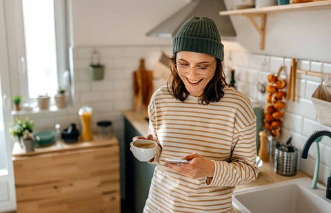 xDame står på kjøkkenet med en kaffekopp og mobiltelefon i hånda