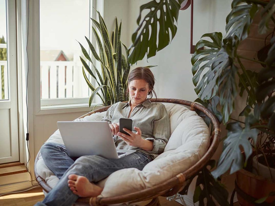 Dame slapper av i stolen med mobil og pc, til illustrasjon for økt boliglånsrente