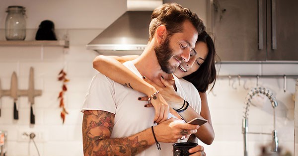 Par står på kjøkkenet og klemmer og ser på mobiltelefonen, til illustrasjon for samboerkontrakt