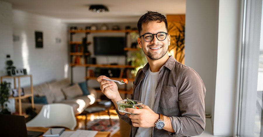 Mann står i stua og smiler mens han spiser en salat, til illustrasjon for refinansiere boliglånet