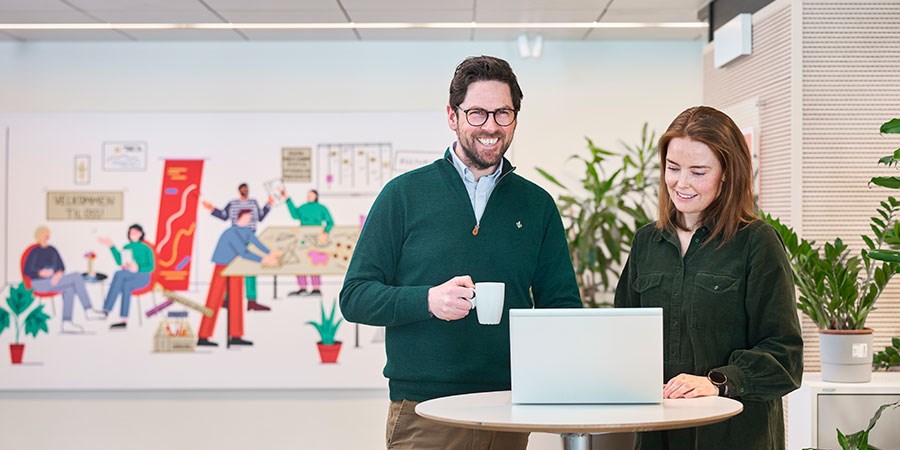 Tom og Siri er boliglånsrådgivere i BN Bank og står i lokalet og smiler med en kaffekopp og PC, til illustrasjon for rentekalkulator