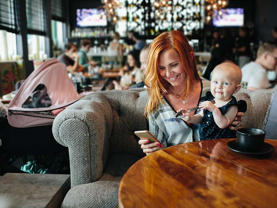 Kvinne sitter på restaurant med et barn på fanget og mobilen i hånda, til illustrasjon for budrunde