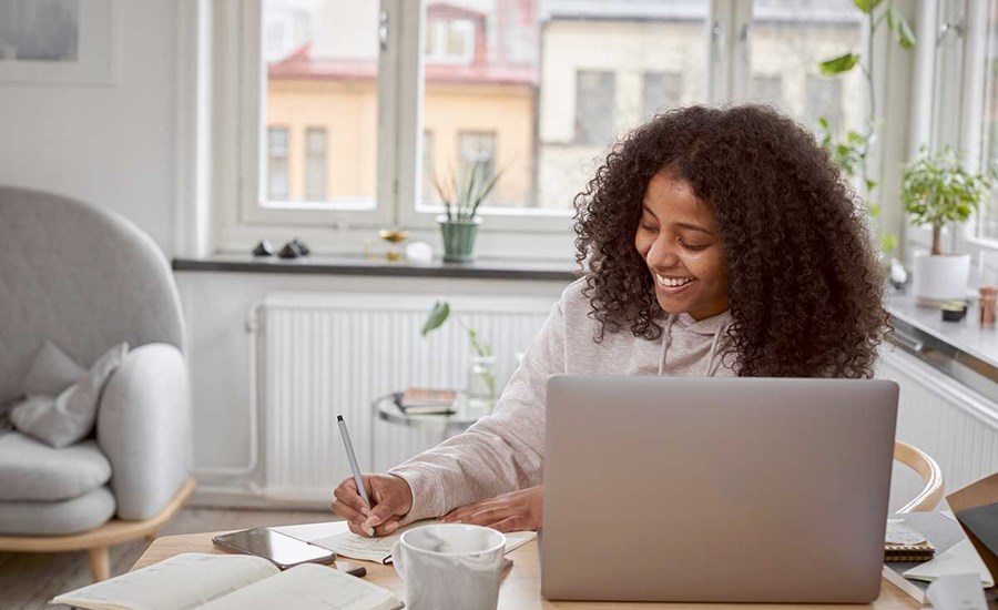 Dame sitter på spisebord og skriver notater med en PC fremfor seg, til illustrasjon for refinansiere forbruksgjeld